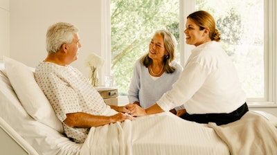 Male patient in a hospital bed with a female family member sitting on the edge of the patient bed and another female family member sitting on a chair next to the bed.