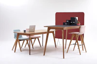 A pink naughtone Viv Wood Stool is stationed at a gray Dalby Bar Height Table. Two Viv Wood Chairs are also stationed at another Dalby Table.