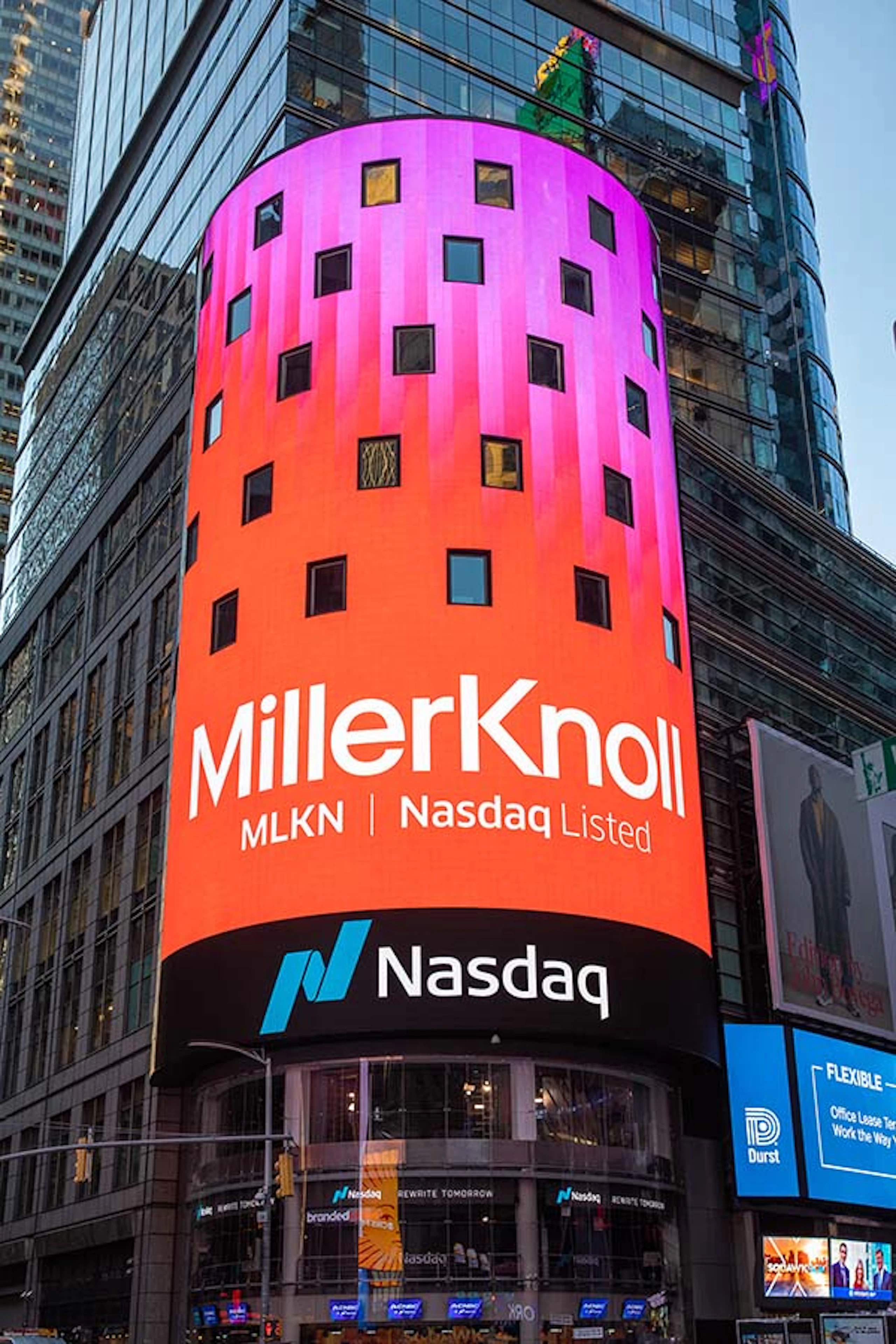 A brightly lit pink and red billboard on Wall Street advertising the merger of MillerKnoll. 