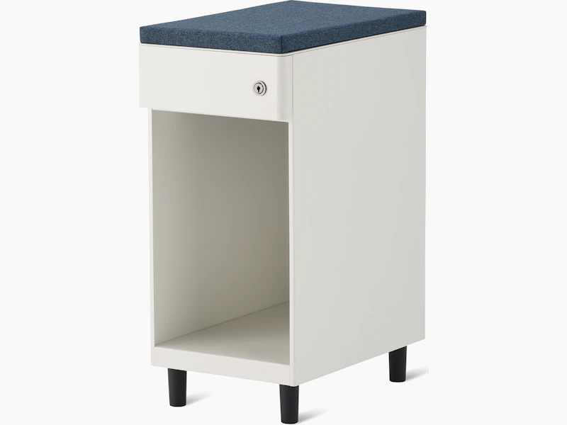 Slim, white storage pedestal on black feet with a drawer and a lock, a blue cushion top, and opening below drawer for storage.