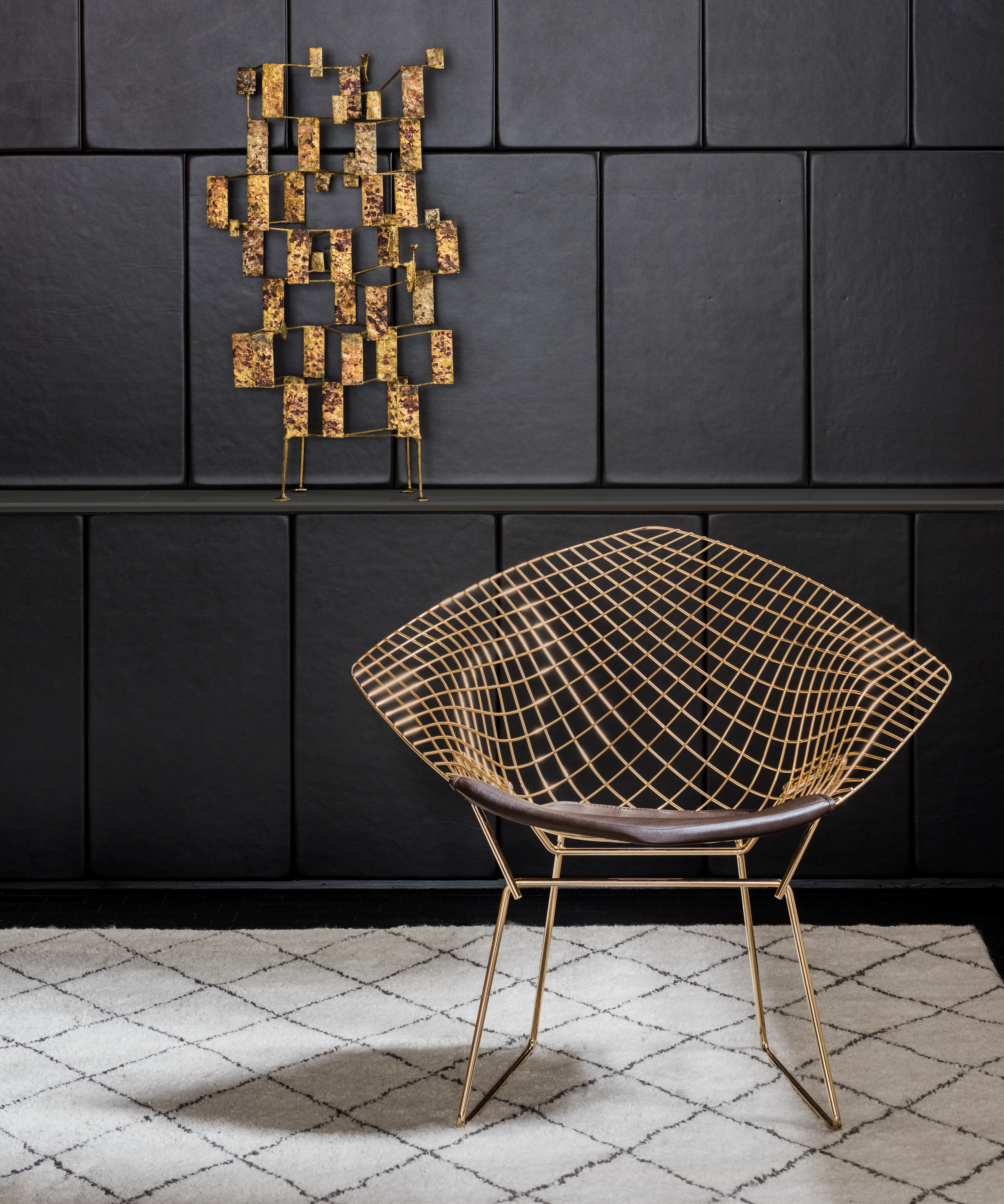 A Bertoia Diamond Chair sitting in front of a black-tiled wall with a piece of geometric gold artwork on it.