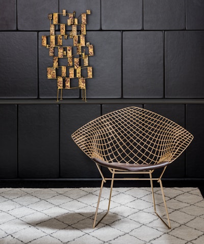 A Bertoia Diamond Chair sitting in front of a black-tiled wall with a piece of geometric gold artwork on it.