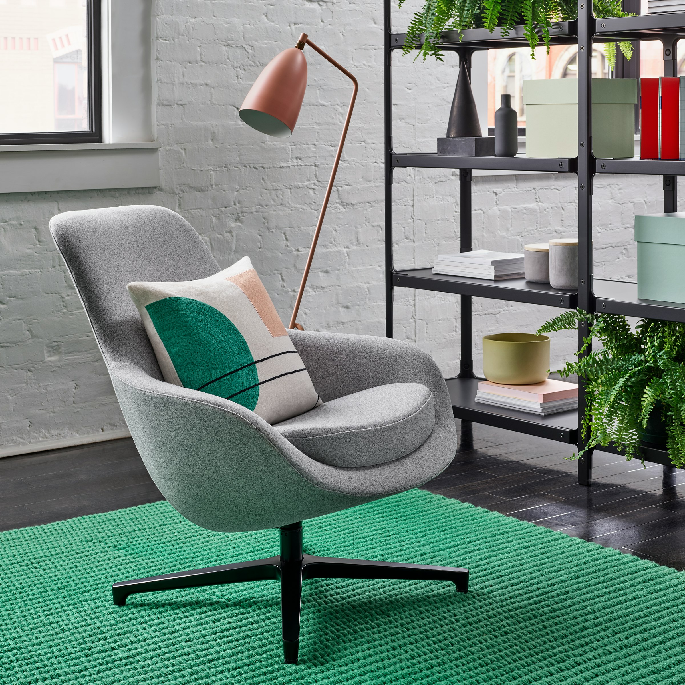 A light grey Saiba Lounge Chair with a black base on a green rug in front of a black Steelwood Shelving System and a salmon Grasshopper Floor Lamp. The scene is set in front of a white wall with windows.