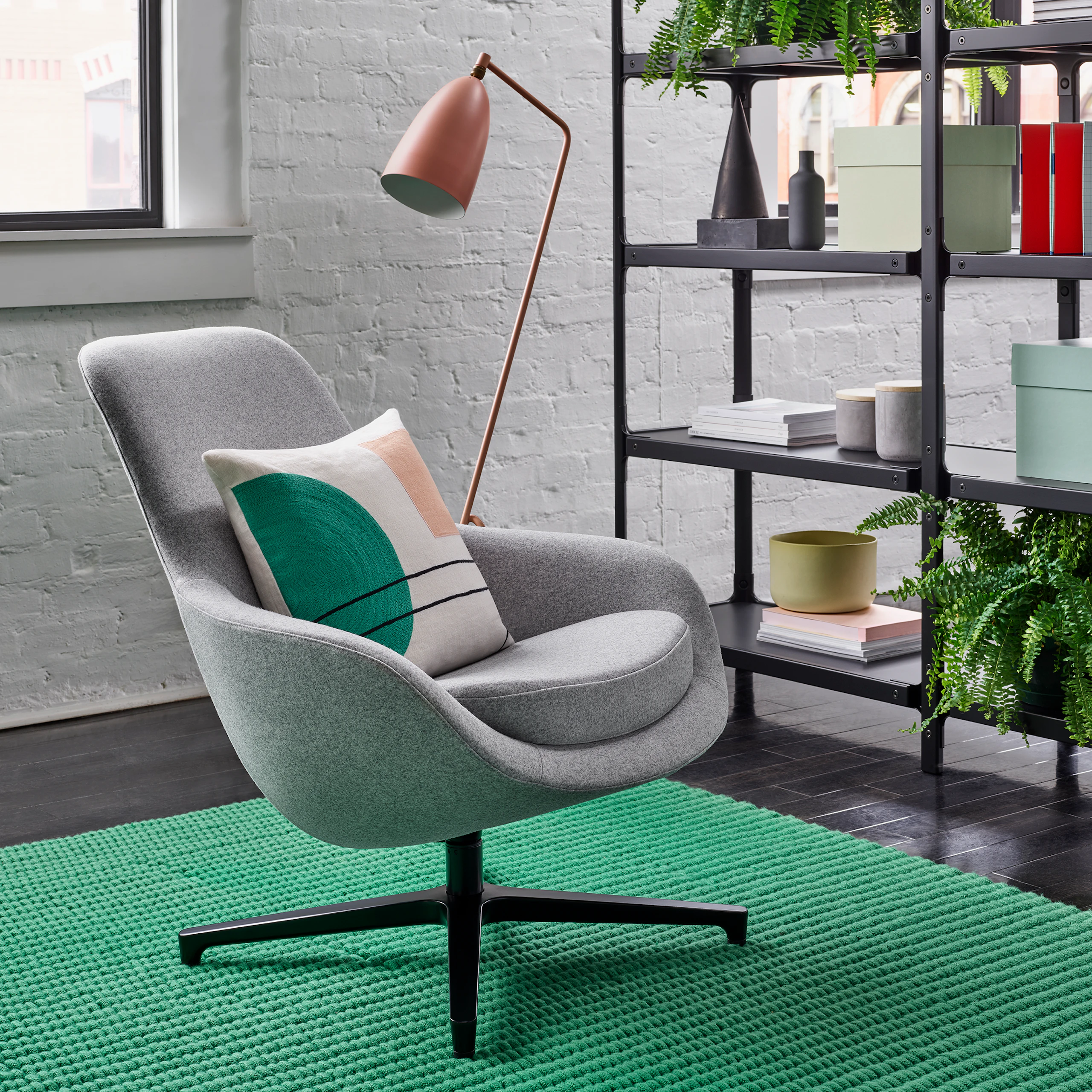 A light grey Saiba Lounge Chair with a black base on a green rug in front of a black Steelwood Shelving System and a salmon Grasshopper Floor Lamp. The scene is set in front of a white wall with windows.