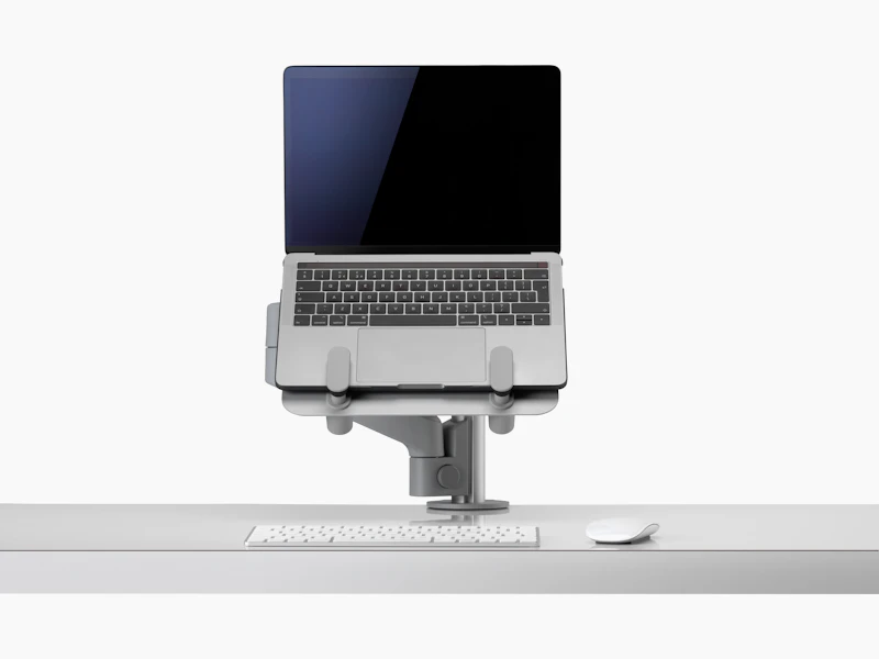 An open laptop raised from a desk and supported by a grey Lima Laptop Mount and Lima Monitor Arm.