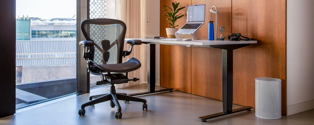 Home office set up showing a height adjustable desk and black office chair positioned next to a window.