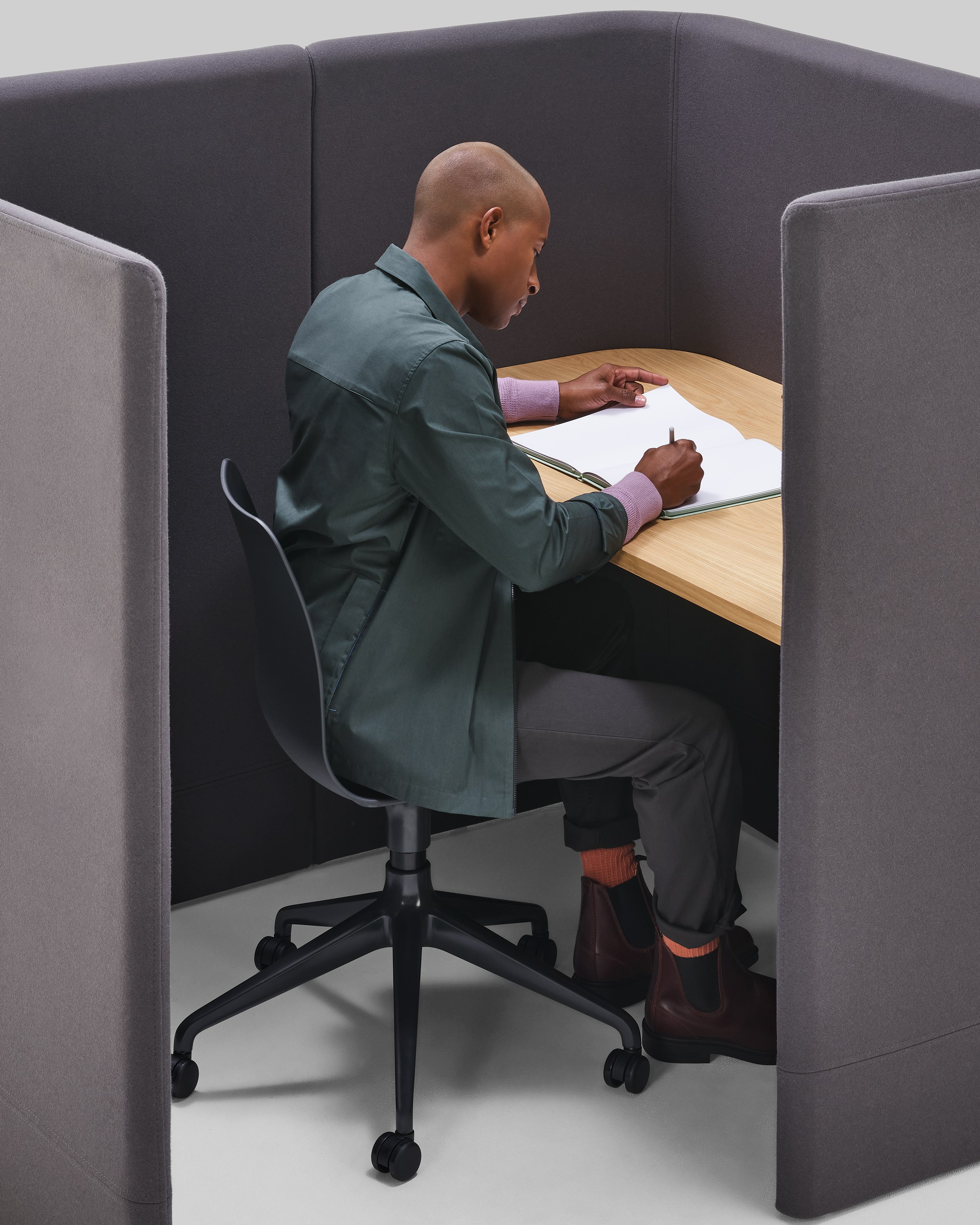 Close up of a man sitting on a Polly chair, writing within a grey Pullman Desk Pod.