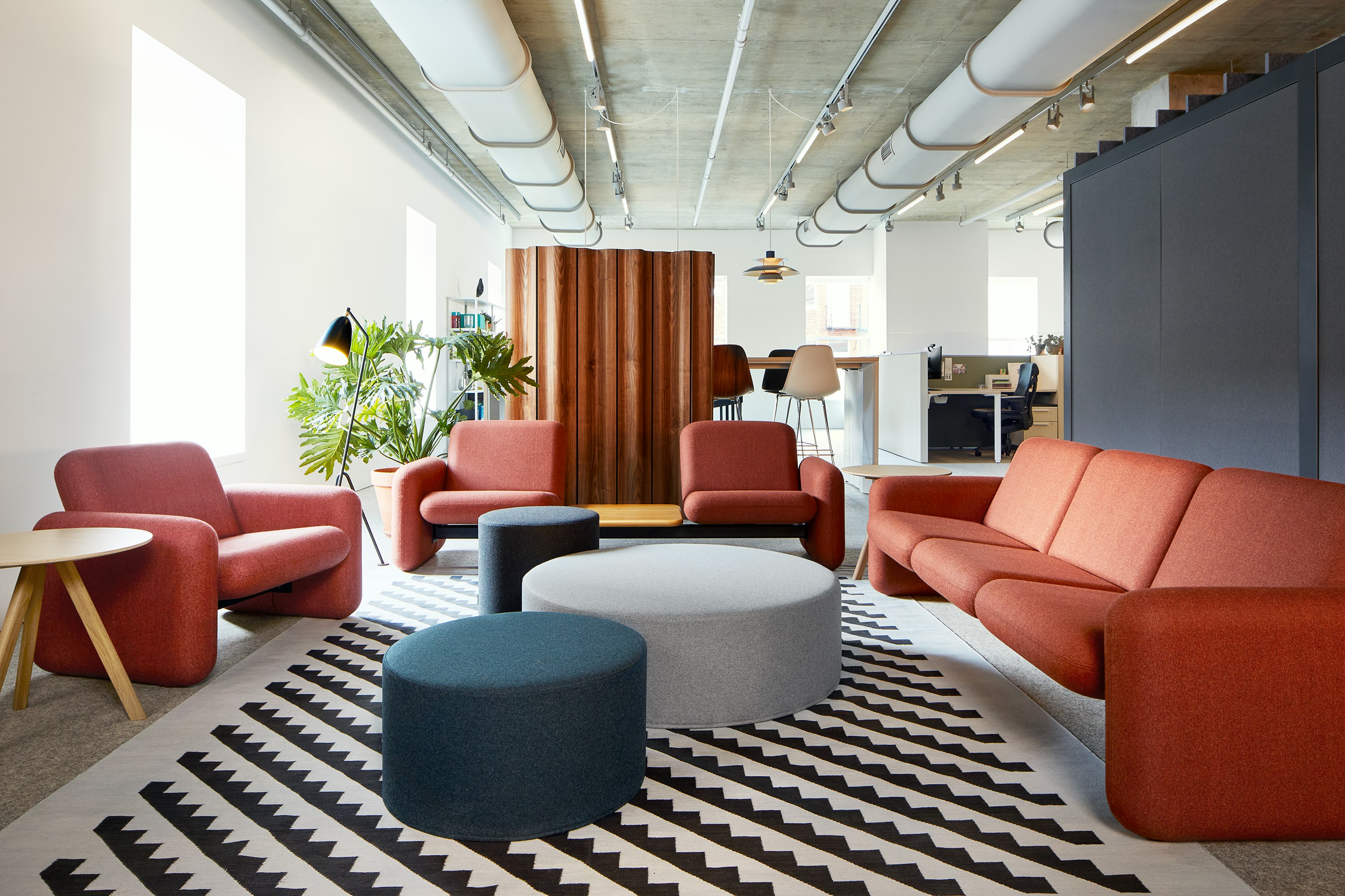 A Wilkes Modular Sofa Group Chair, Two Seat Sofa with white oak table and Three Seat Sofa all in orange-brown surround three poufs in blue and grey and a patterned rug.