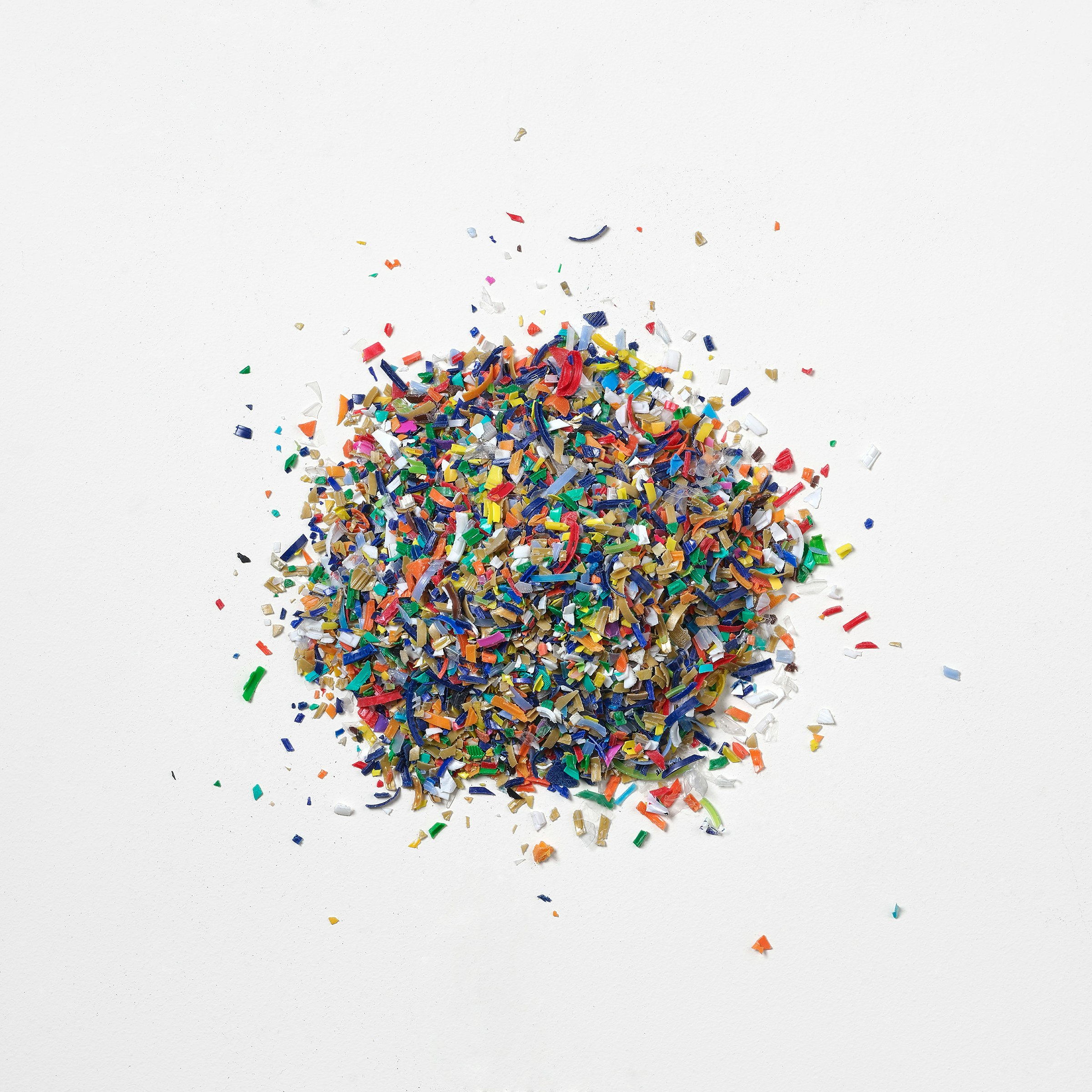 A round pile and scattered bits of multi-colored recycled ocean-bound plastic flakes on a white background.