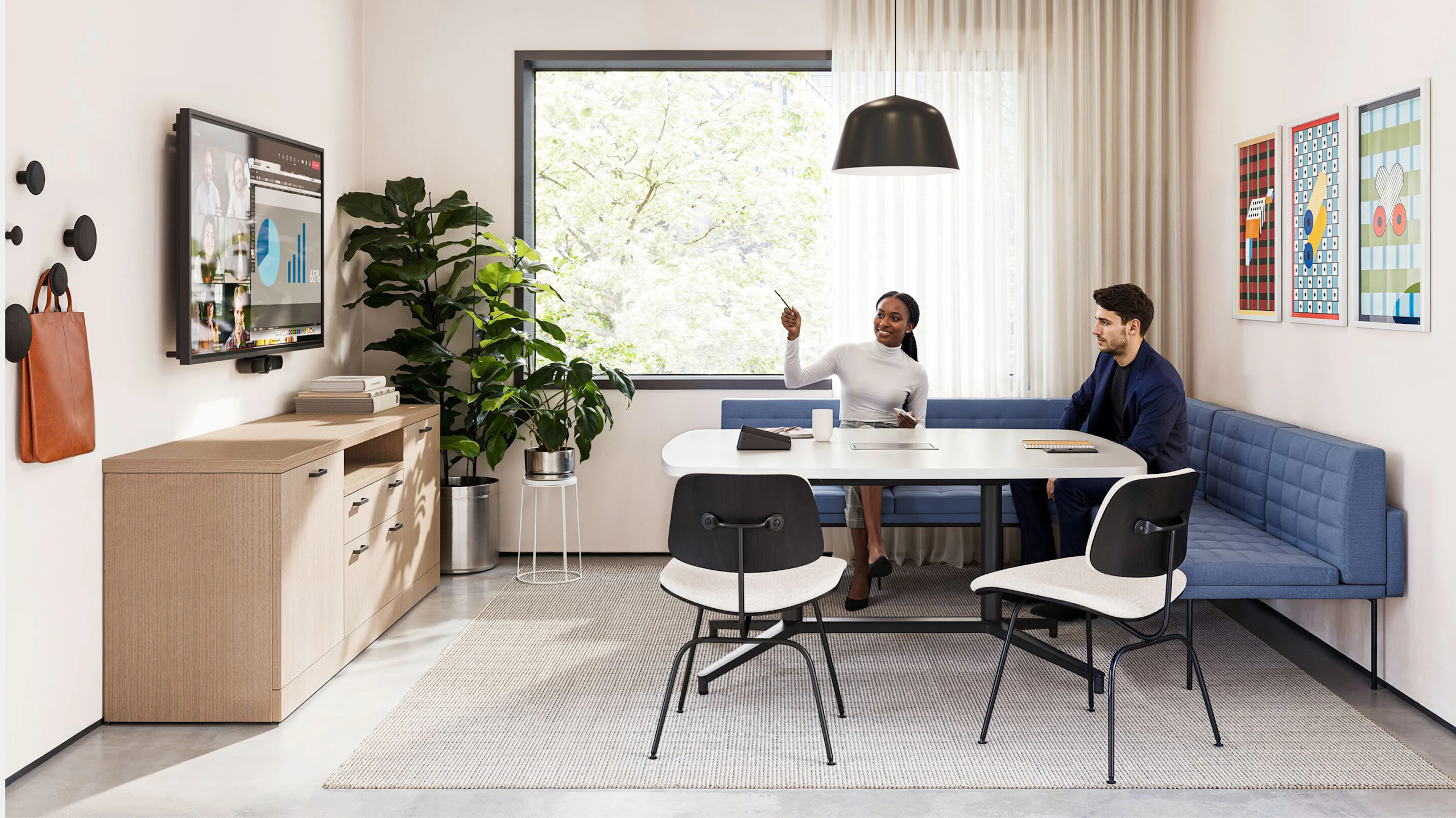 A group setting featuring two people sitting on a blue corner bench while participating in a virtual meeting in a conference room furnished with Geiger Tuxedo Component Lounge seating, a Knoll Grasshopper Table, and a Knoll Florence Knoll Credenza