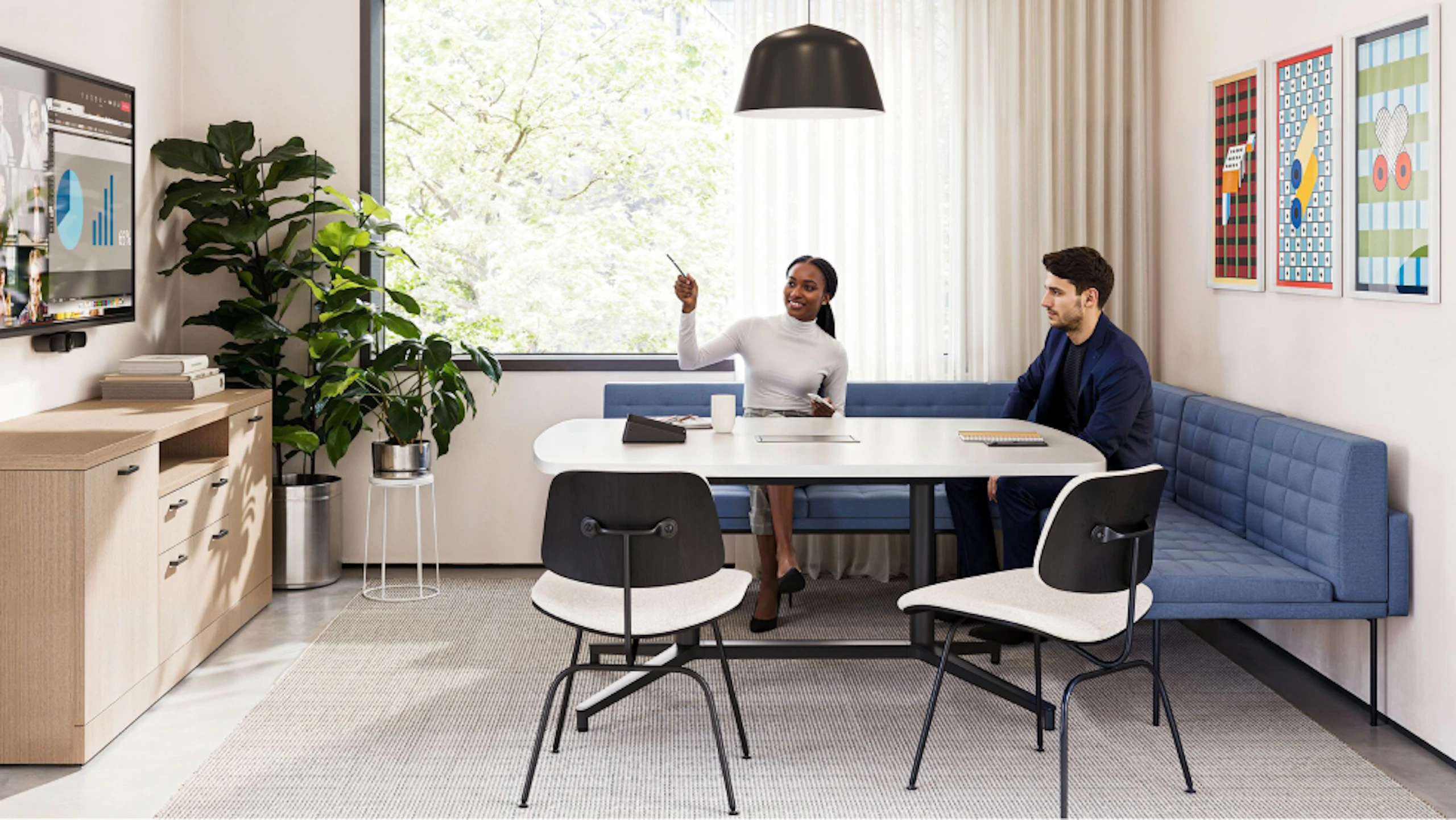 A group setting featuring two people sitting on a blue corner bench while participating in a virtual meeting in a conference room furnished with Geiger Tuxedo Component Lounge seating, a Knoll Grasshopper Table, and a Knoll Florence Knoll Credenza