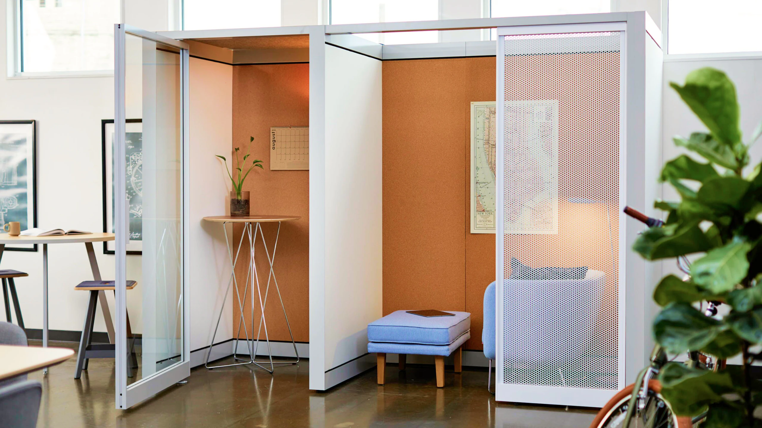 Green foliage next to an orange Knoll Rockwell Unscripted Creative Wall refuge room furnished with a grey Rockwell Unscripted Petite Club Chair and Ottoman and a counter height table.