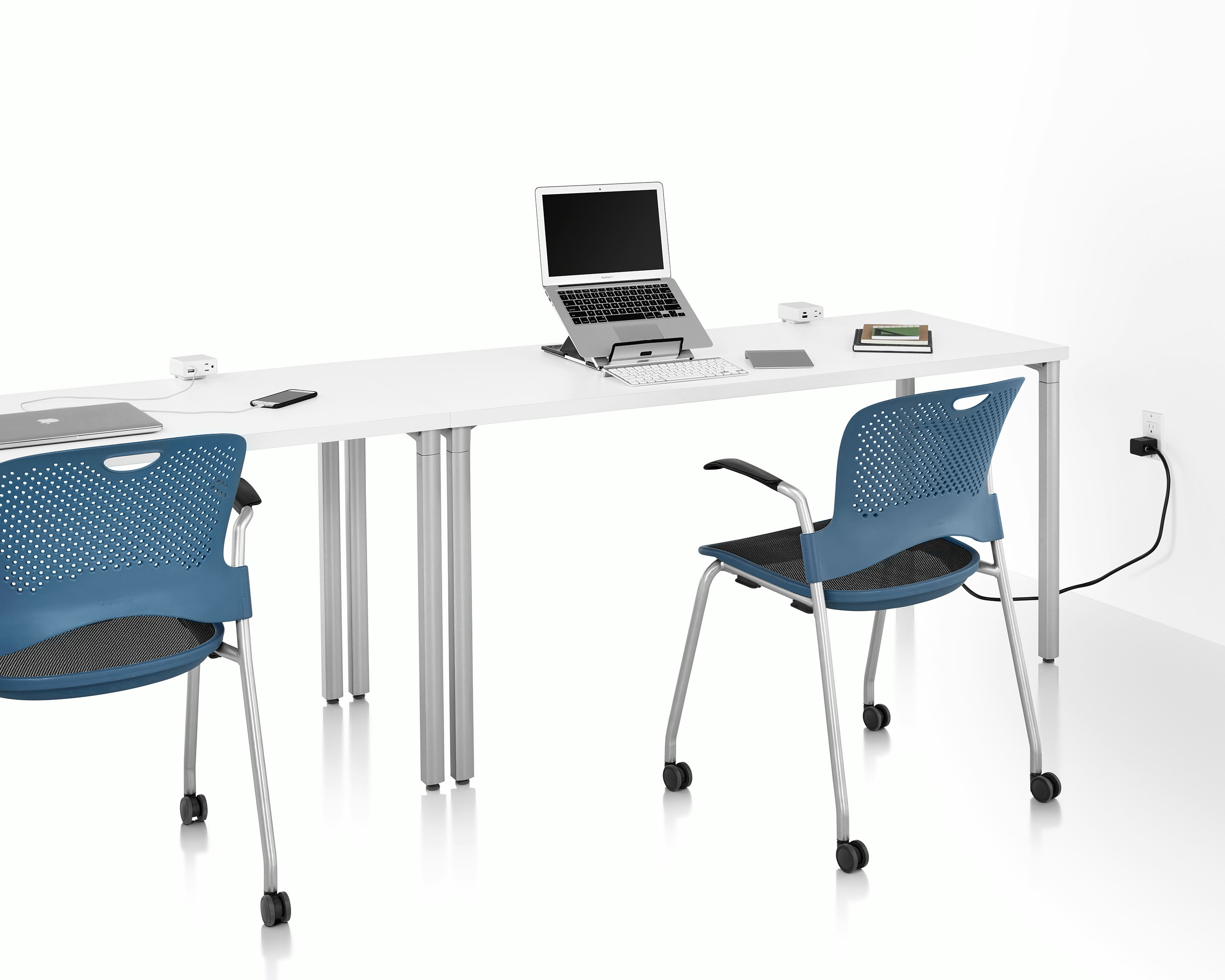 Two white tables and two blue chairs on casters with power access sources that include USB-A port, USB-C port, and A/C power outlet. Laptop, keyboard, phone, and books are also on top of table