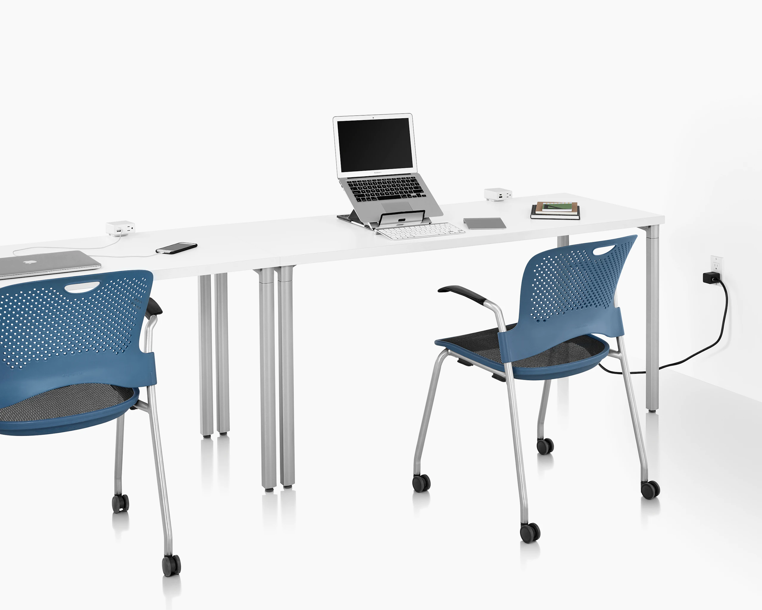 Two white tables and two blue chairs on casters with power access sources that include USB-A port, USB-C port, and A/C power outlet. Laptop, keyboard, phone, and books are also on top of table