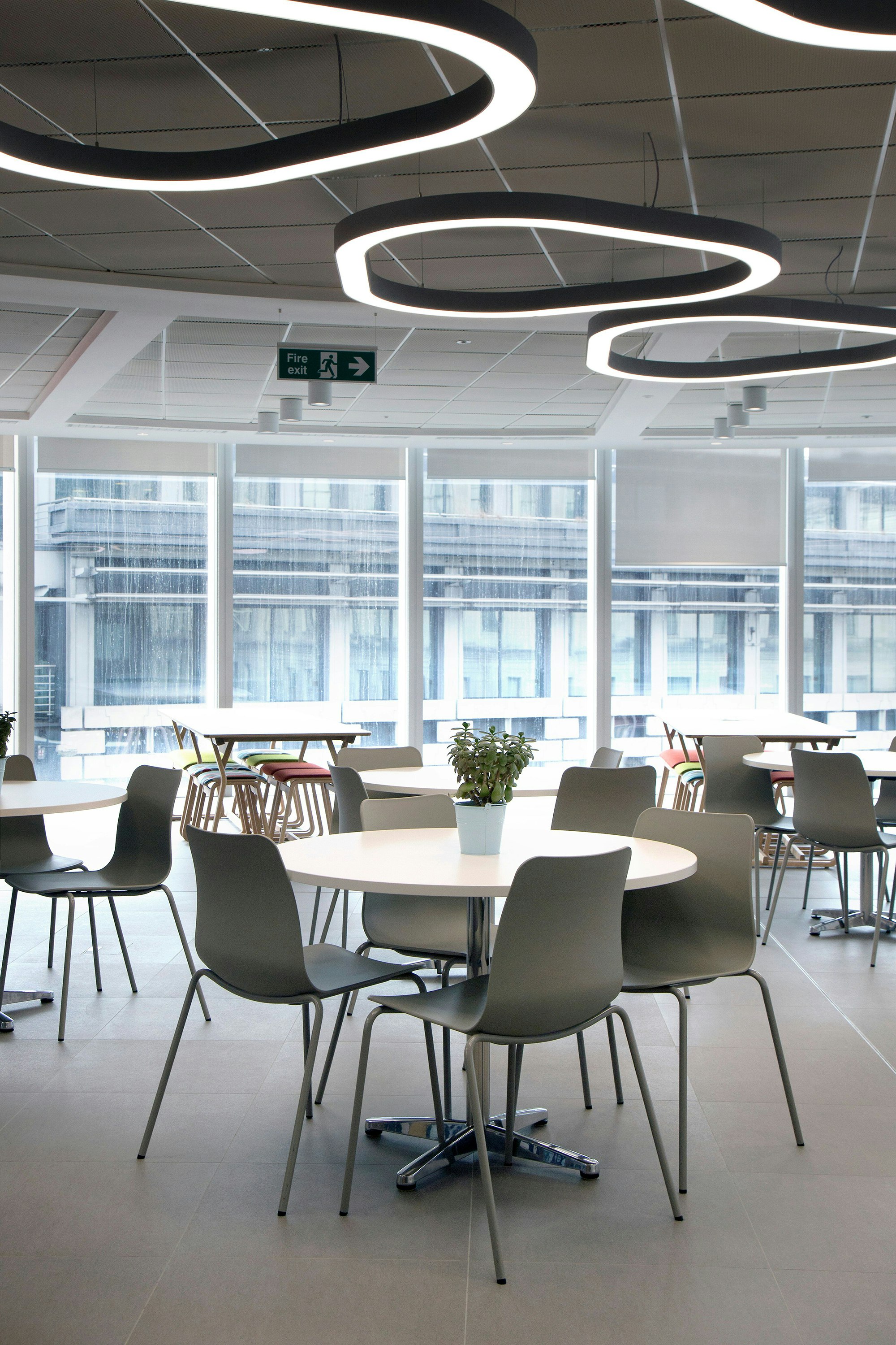 Four gray Polly Side Chairs sit around a white tabletop with a metal base in a dining hall setting.