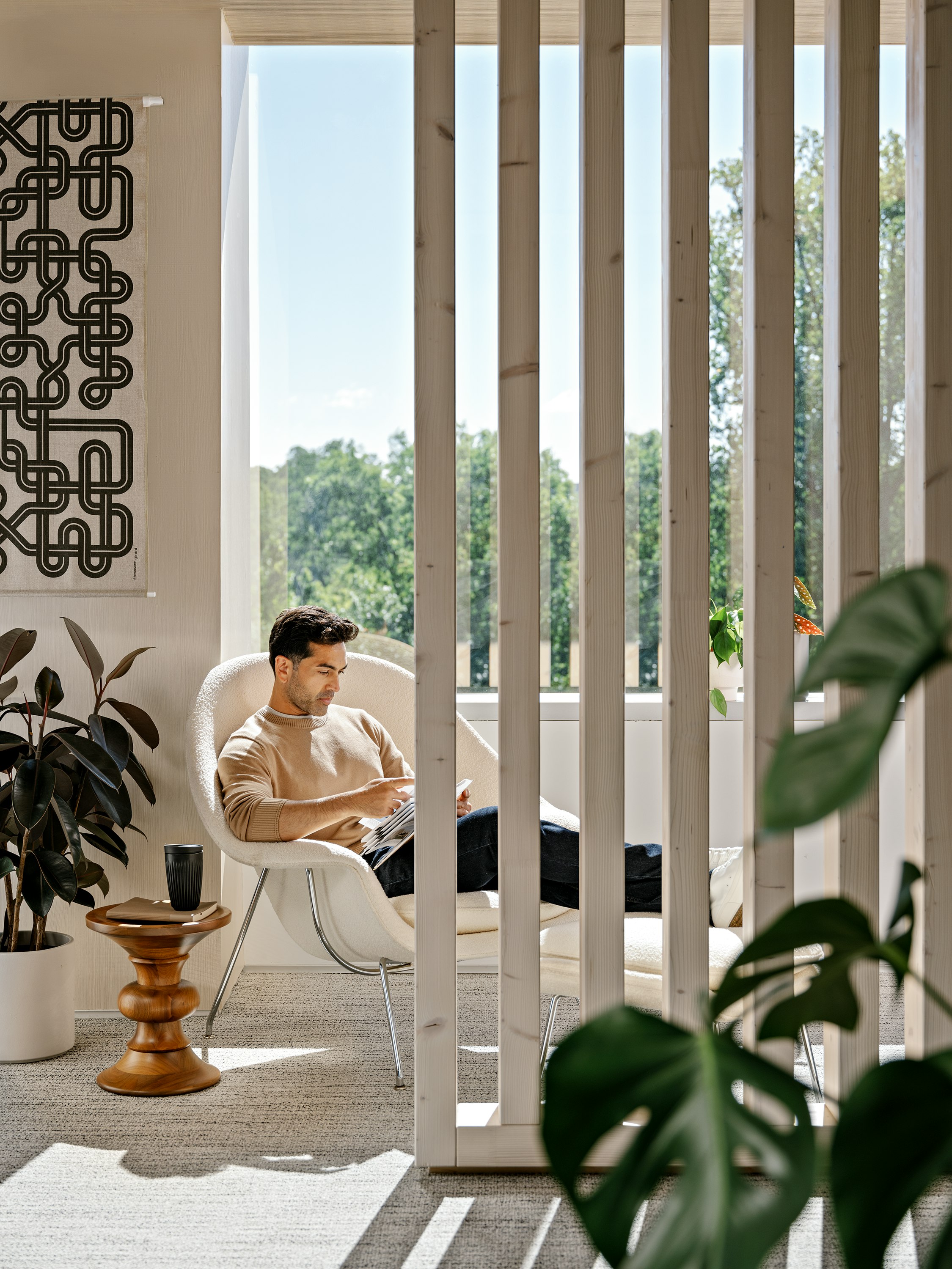 A person with short brown hair, wearing a beige sweater and blue jeans, sitting in a white Knoll Womb Chair and Ottoman while reading next to green potted plants