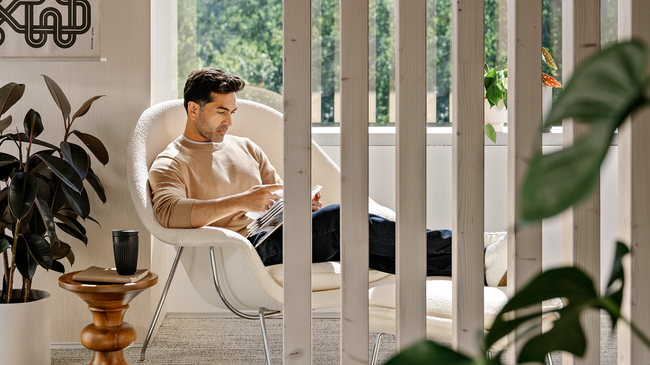 A person with short brown hair, wearing a beige sweater and blue jeans, sitting in a white Knoll Womb Chair and Ottoman while reading next to green potted plants