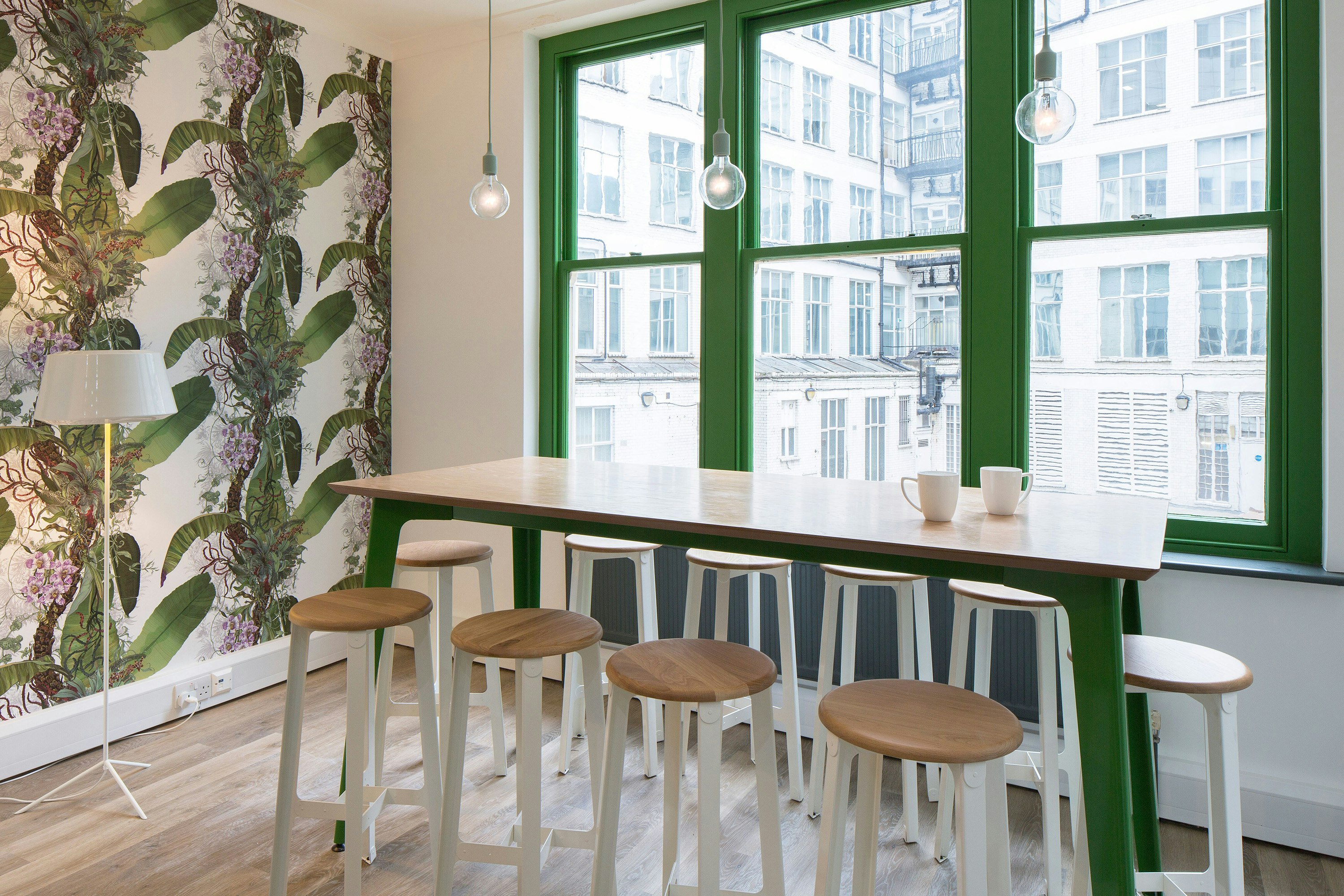 A naughtone Fold Bar Height Table with a wood veneer top and a green base in front of a window surrounded by white Construct Stools.