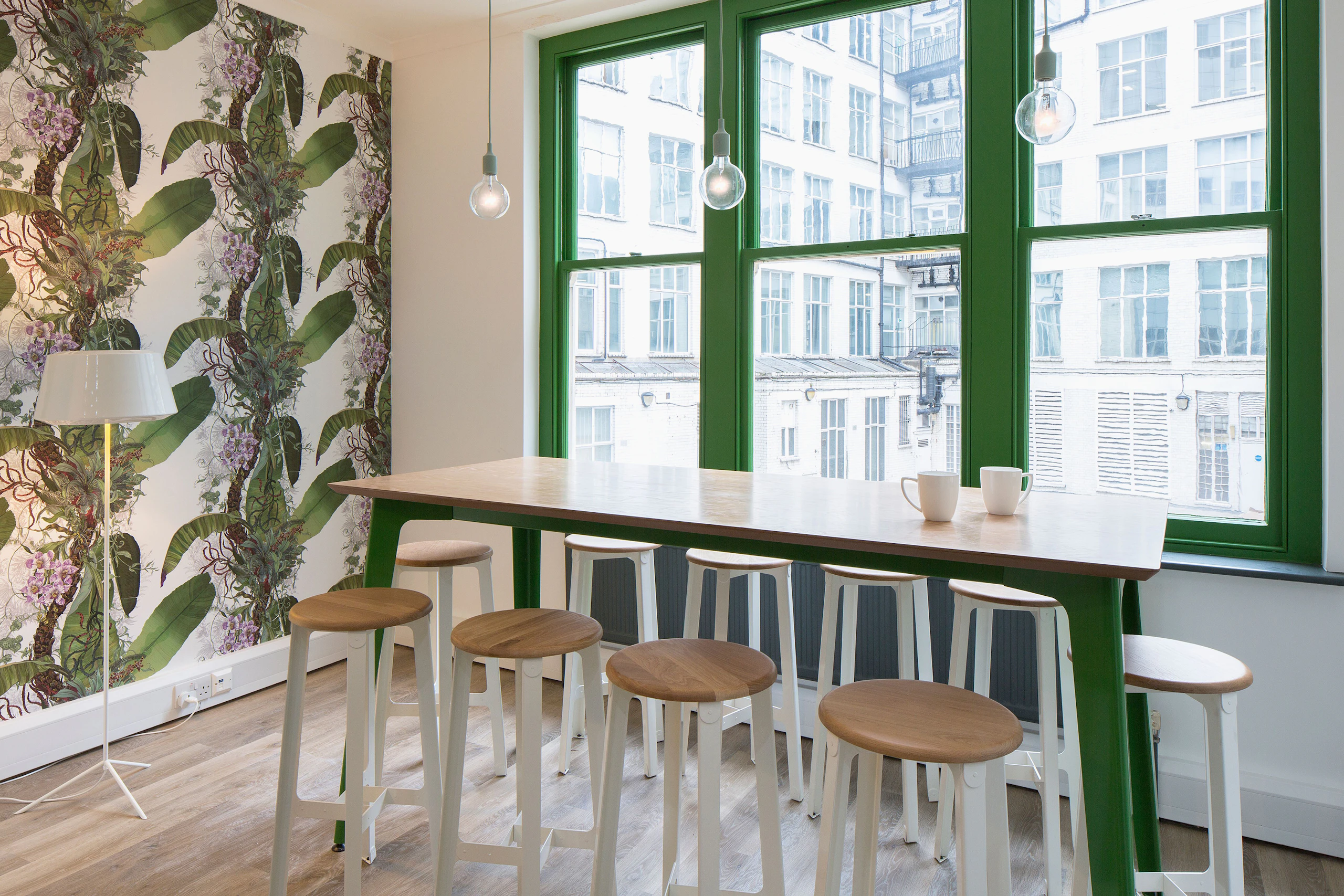 A naughtone Fold Bar Height Table with a wood veneer top and a green base in front of a window surrounded by white Construct Stools.