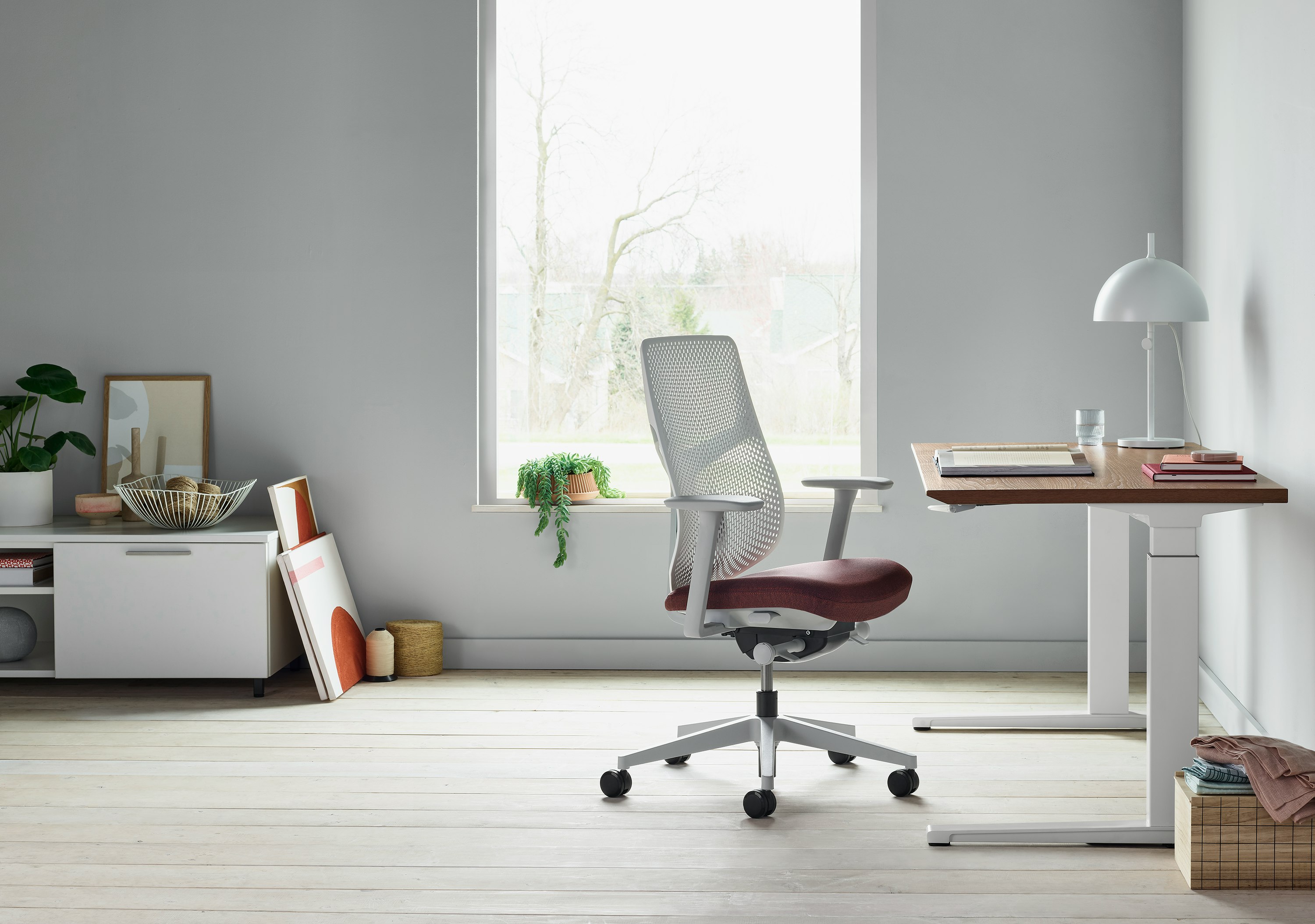 A white Verus Chair with dark red seat next to a Renew Sit-to-Stand Table with a brown surface close to a window.