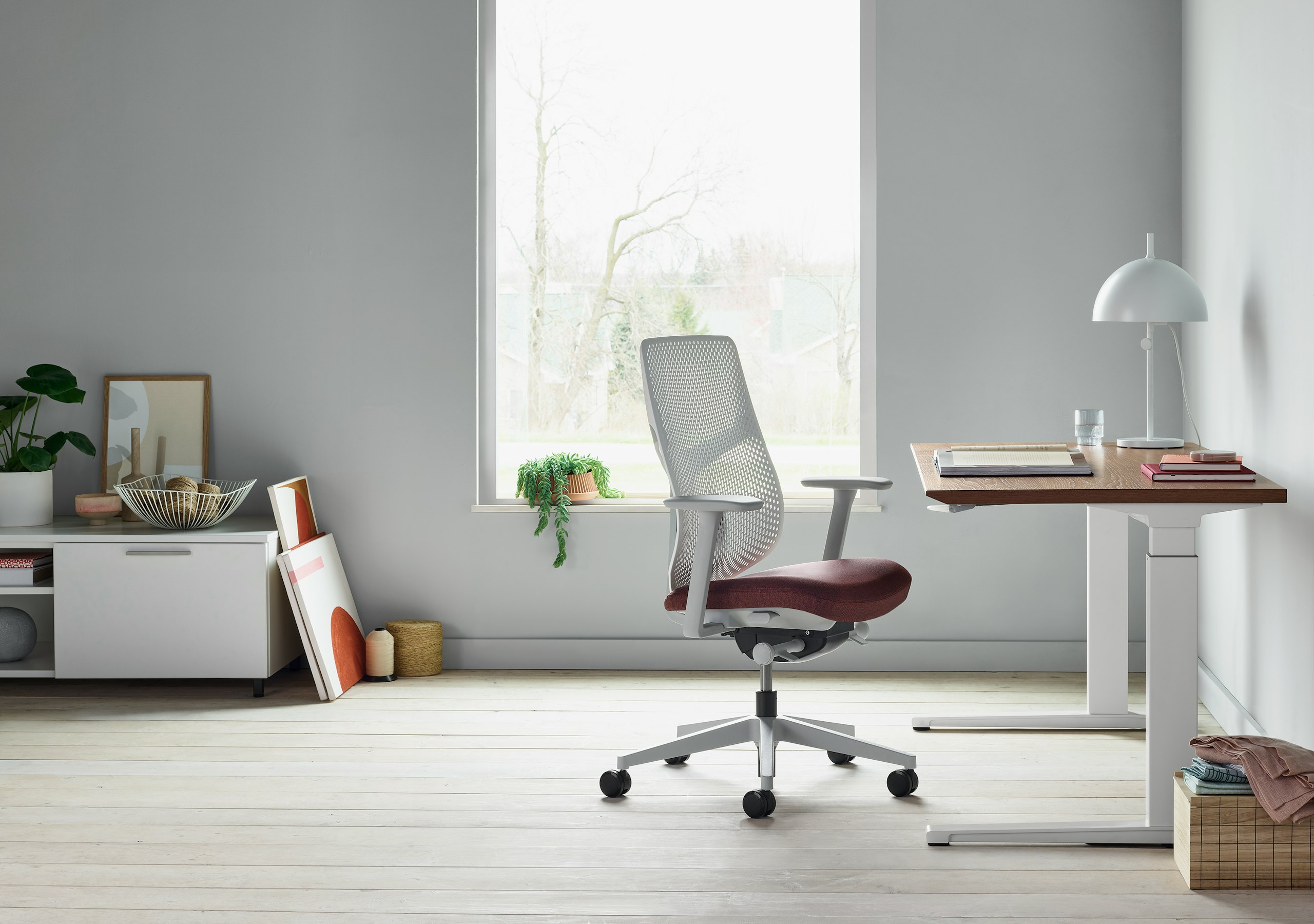 A white Verus Chair with dark red seat next to a Renew Sit-to-Stand Table with a brown surface close to a window.