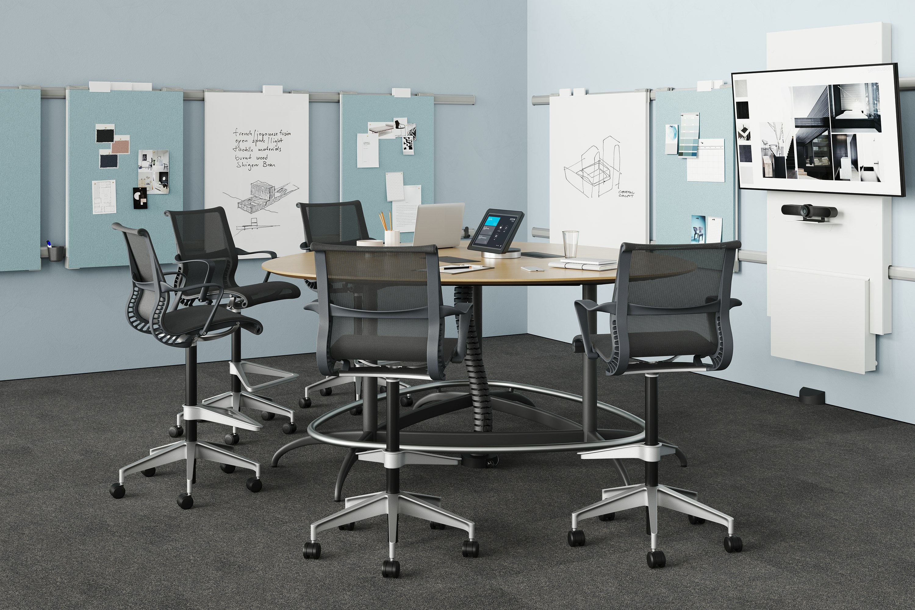 A meeting room setting featuring an Exclave table with five black Setu Stools in front of a monitor and Exclave display elements.