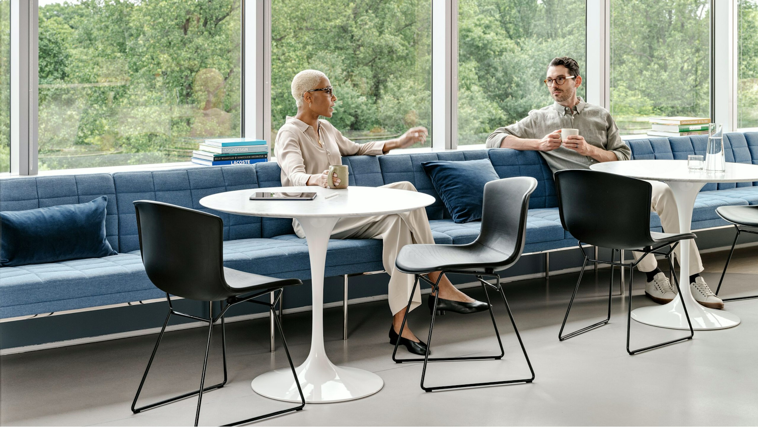 Dos personas conversan en una zona de estar compartida amueblada con mesas Saarinen, sillas Knoll Bertoia y un banco azul.