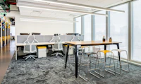 A black naughtone Fold Bar Height Table and stools in a high-rise office, a benching system and Sayl Chairs by Herman Miller in the background.