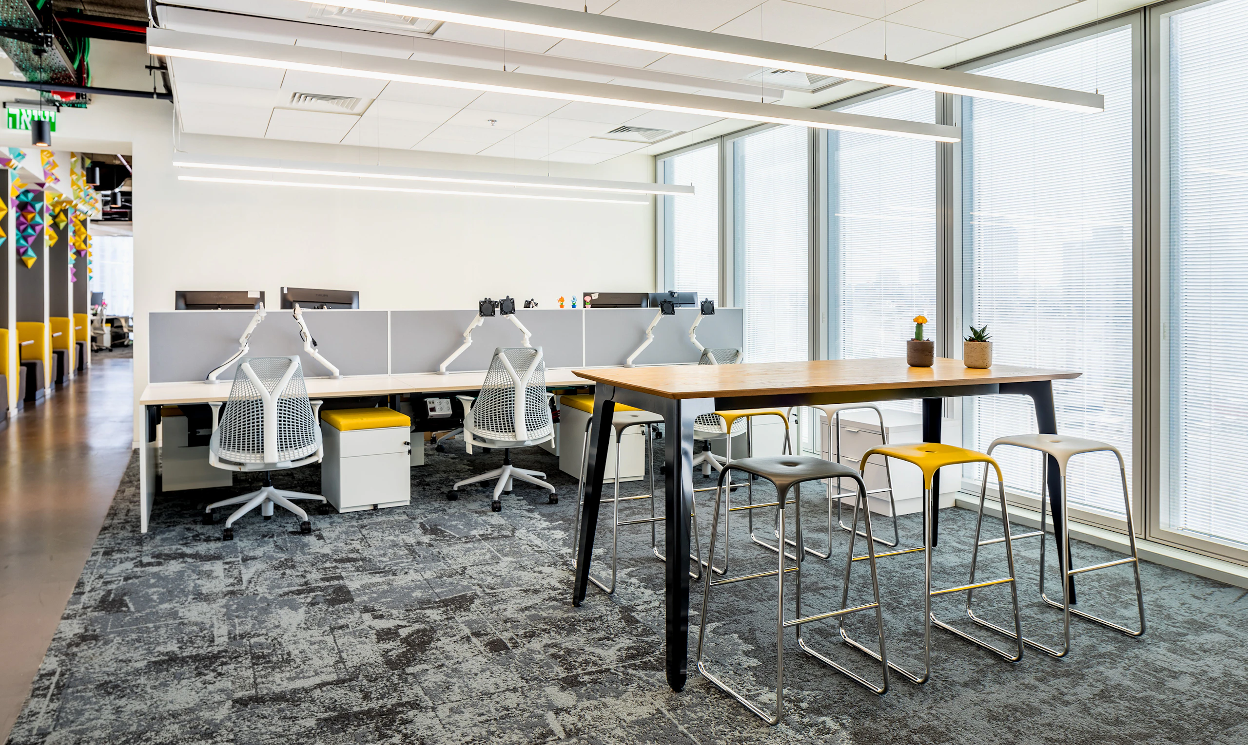 A black naughtone Fold Bar Height Table and stools in a high-rise office, a benching system and Sayl Chairs by Herman Miller in the background.