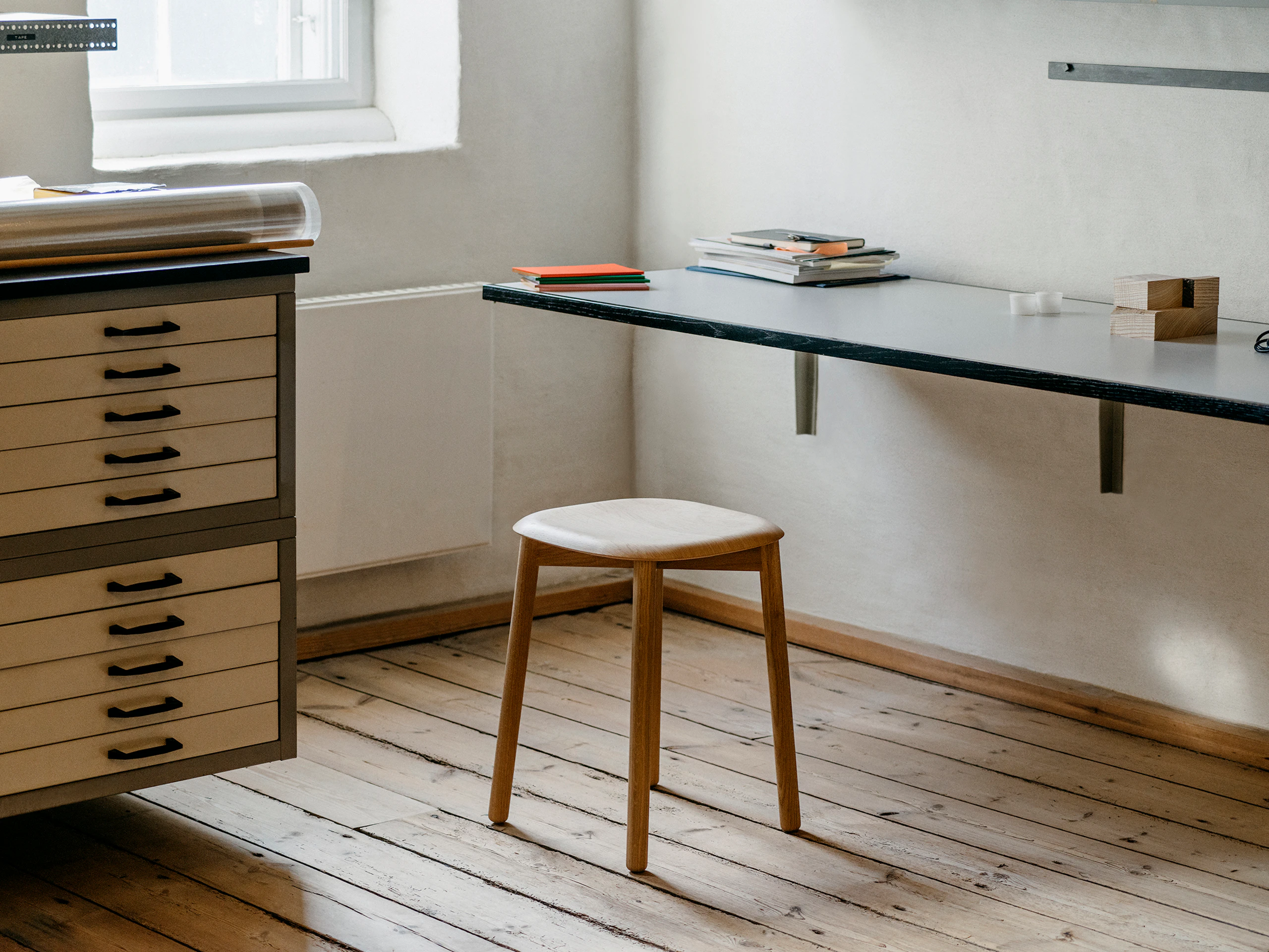 A Soft Edge wood stool in Oak beside a counter and a stack of drawers.