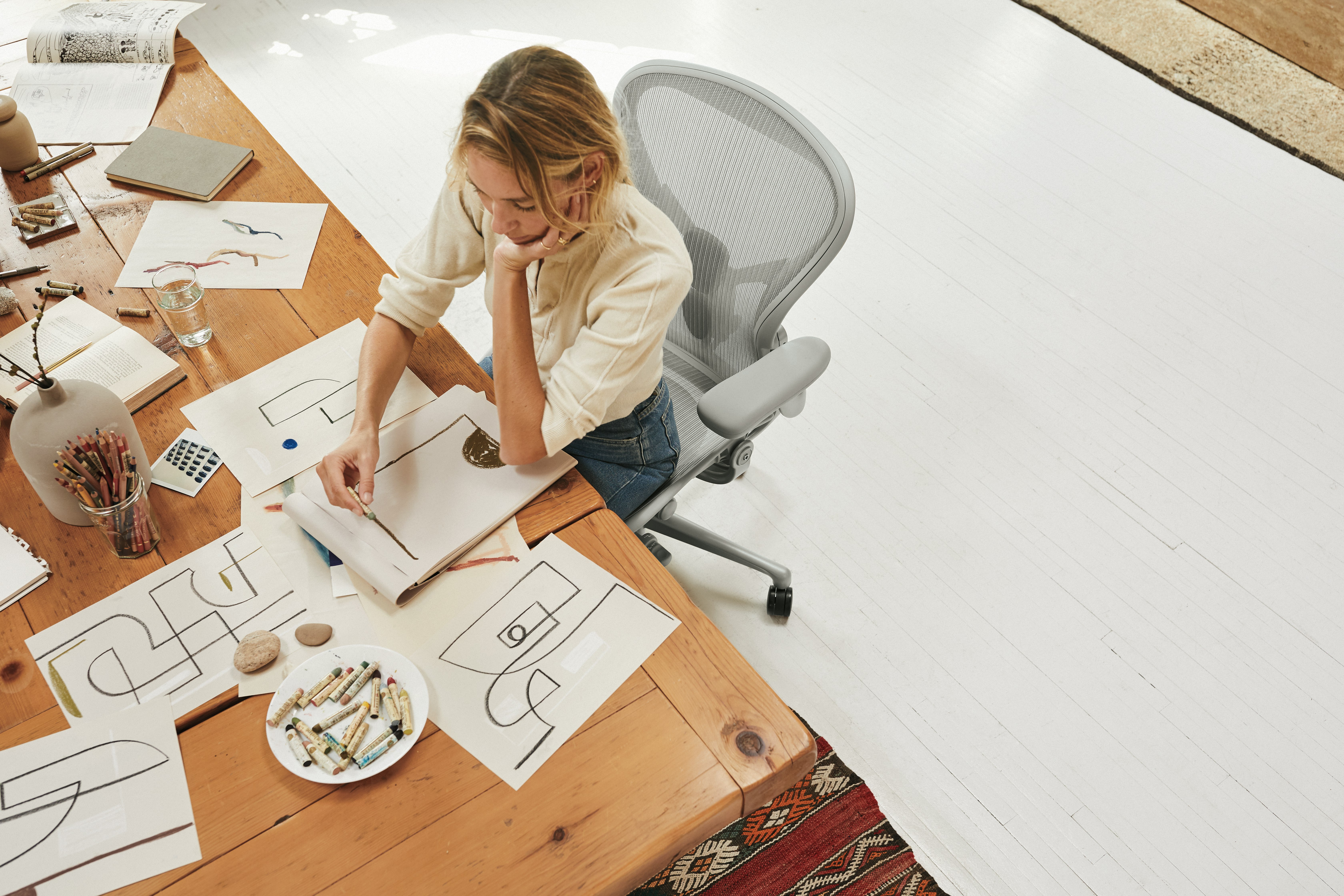A woman working on a drawing while sitting in a mineral Aeron Chair at a dining room table with a medium tone wood top.