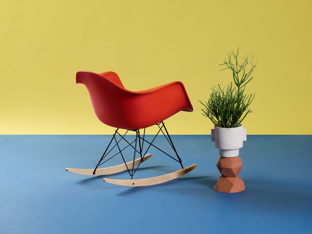 Single red Eames Molded Plastic Rocking Armchair with yellow background styled with plant.