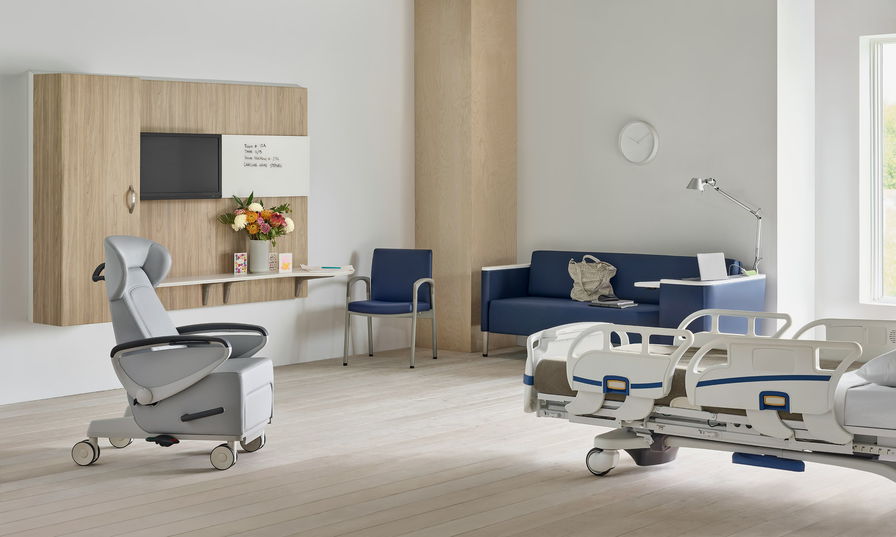 Compass System footwall in a patient room with a gray Ava recliner, blue Palisade Flop Sofa, a blue Valor Side Chair and a hospital bed.