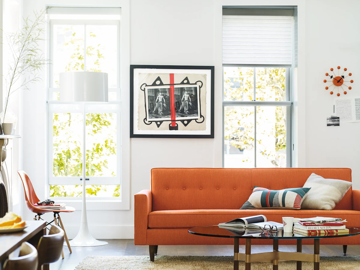 Bright living room with orange sofa, framed wall art, and round glass coffee table.