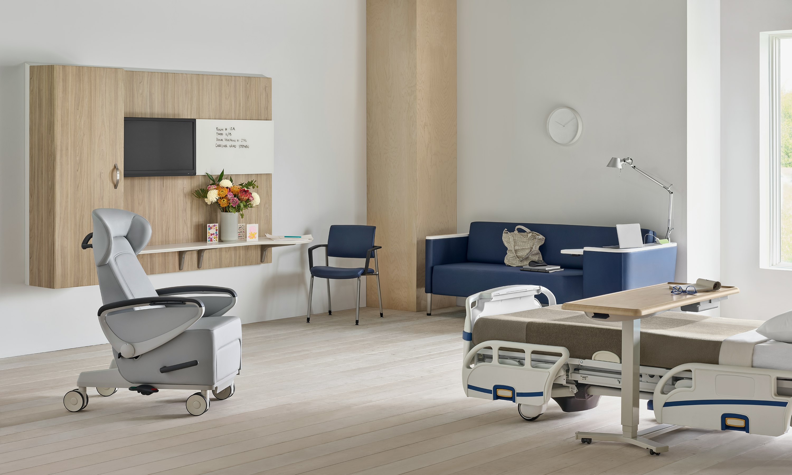 Compass System footwall in a patient room with a gray Ava patient recliner, a blue Palisade Flop Sofa, a fully-upholstered blue Verus side chair, and a Mirage Overbed Table over a hospital bed.
